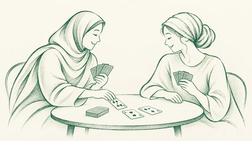 Two smiling women in head coverings playing cards together, one placing a card on the table.